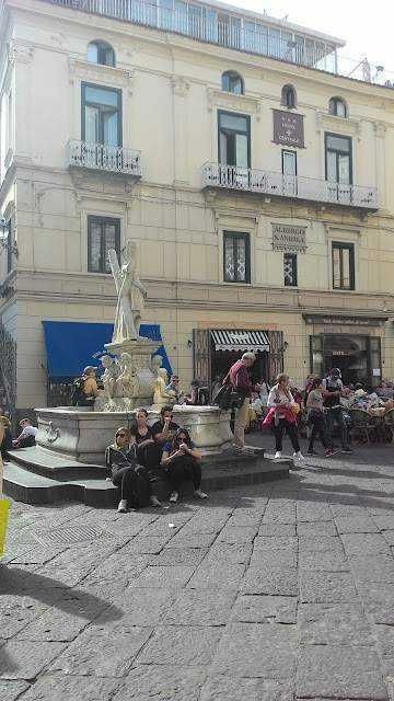 Fontana storica al centro di una piazza affollata con persone sedute e camminando, edificio storico sullo sfondo, atmosfera vivace e traffico pedonale, tipica scena urbana di una città italiana.