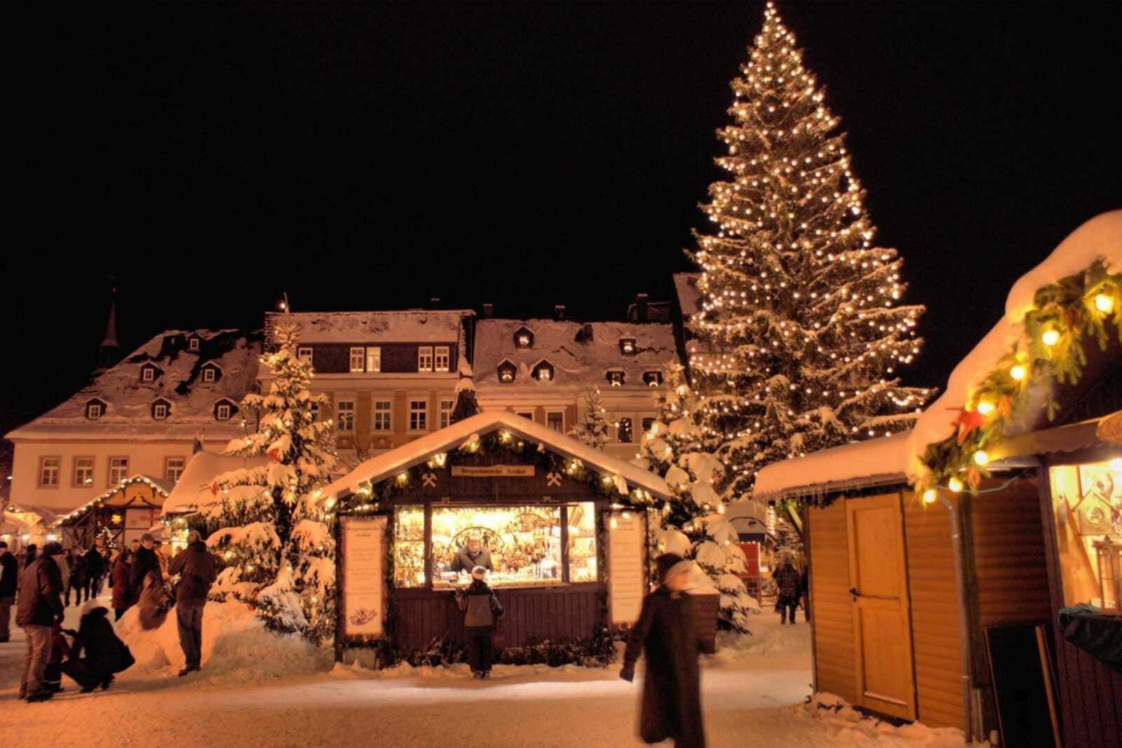 Baite di legno con luci natalizie, grandi alberi decorati e mercato di Natale innevato in Montagna, atmosfera magica, tradizione, festeggiamenti invernali e commercio di artigianato e prodotti tipici natalizi.