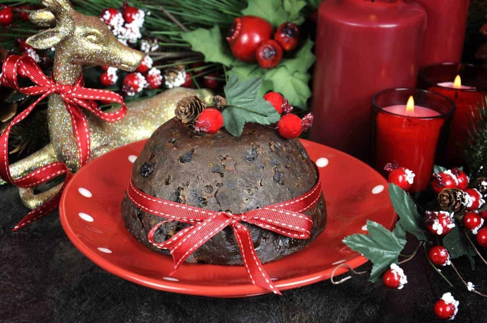Dolce di Natale al panettone con uvetta e cioccolato, decorato con bacche rosse, foglie di agrifoglio e nastri rossi, su un piatto rosso per le festività natalizie.