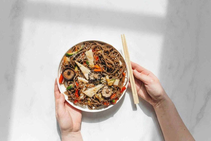 Menu di capodanno: 13 piatti da mettere a tavola 12 Spaghetti di soba con verdure e funghi, piatto giapponese di noodles integrali, servito in una ciotola bianca con bacchette, stile minimalista, presentato su sfondo bianco.