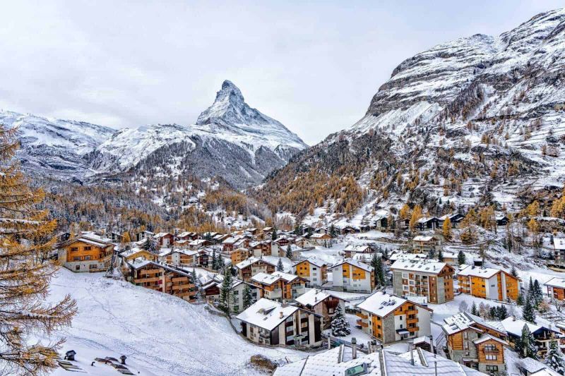 5 destinazioni innevate per il Natale in montagna 7 Montagna innevata e villaggi alpini nel paesaggio invernale, con il famoso Monte Cervino visibile all'orizzonte, durante una giornata nuvolosa, rappresentando le scogliere e le case di montagna.