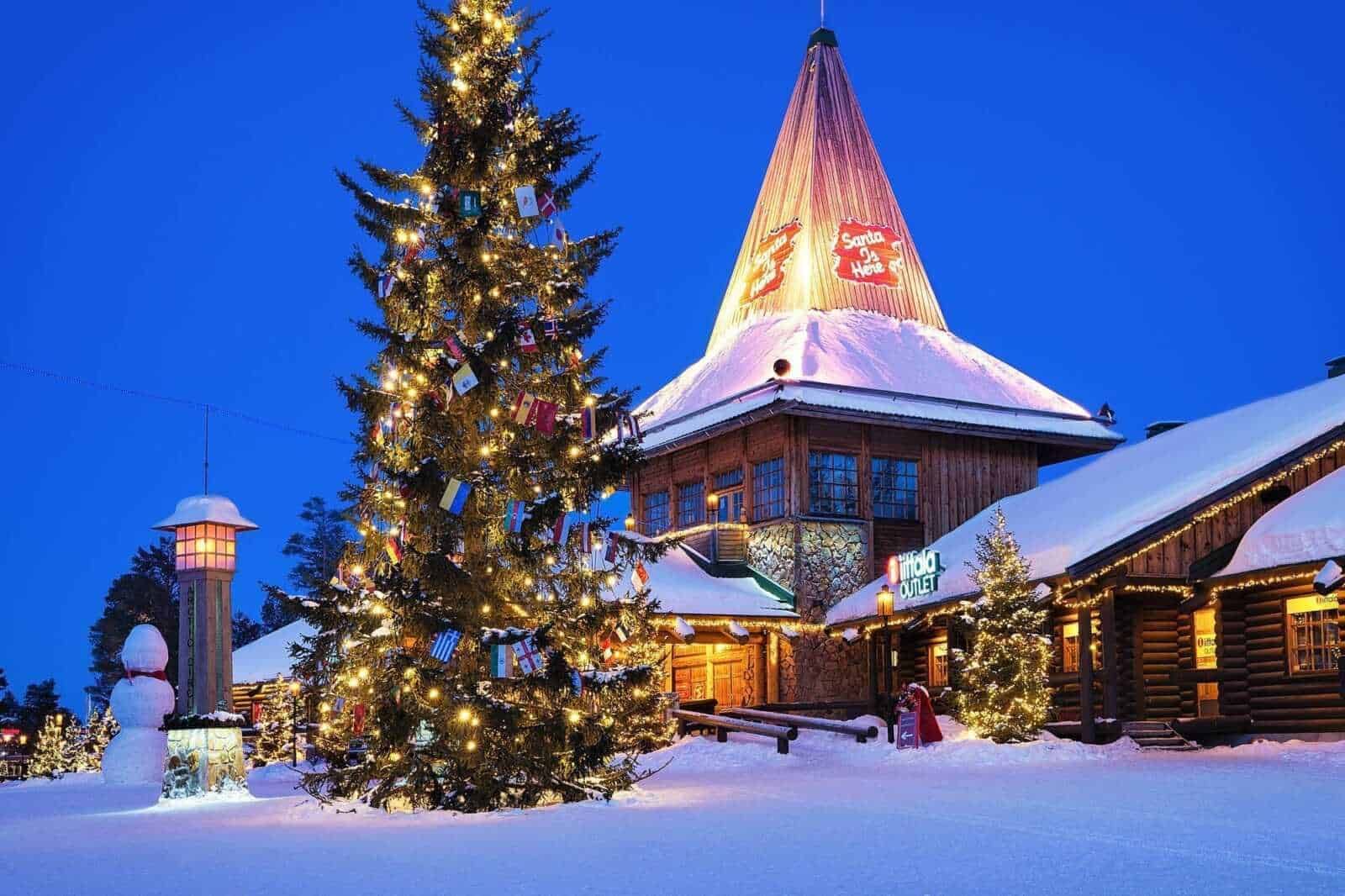 Albero di Natale decorato con luci e regali, atmosfera natalizia all'aperto con neve e chalet in stile alpino, illuminazioni festive e ambiente accogliente, perfetto per il periodo di festa in montagna.