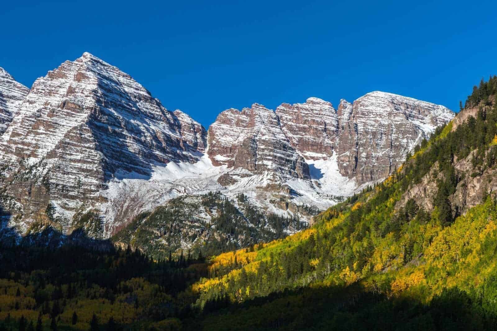 Montagne innevate delle Dolomiti con foresta autunnale sulla valle sottostante, cielo azzurro cristallino, paesaggio naturale italiano.