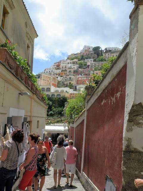 La storia di Positano dalle origini ai giorni nostri 2 Passeggiata tra strette vie di Positano con case colorate sulla collina, panorama di mare e cielo azzurro sullo sfondo, atmosfera mediterranea, turismo, itinerario in Costiera Amalfitana.