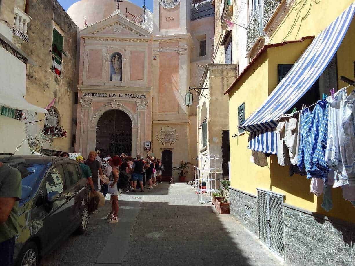Finestra di un vicolo storico a Napoli con persone in fila vicino a una chiesa, barche di lavanderia appese e edifici colorati, espressione dell'atmosfera vivace e caratteristica della città.