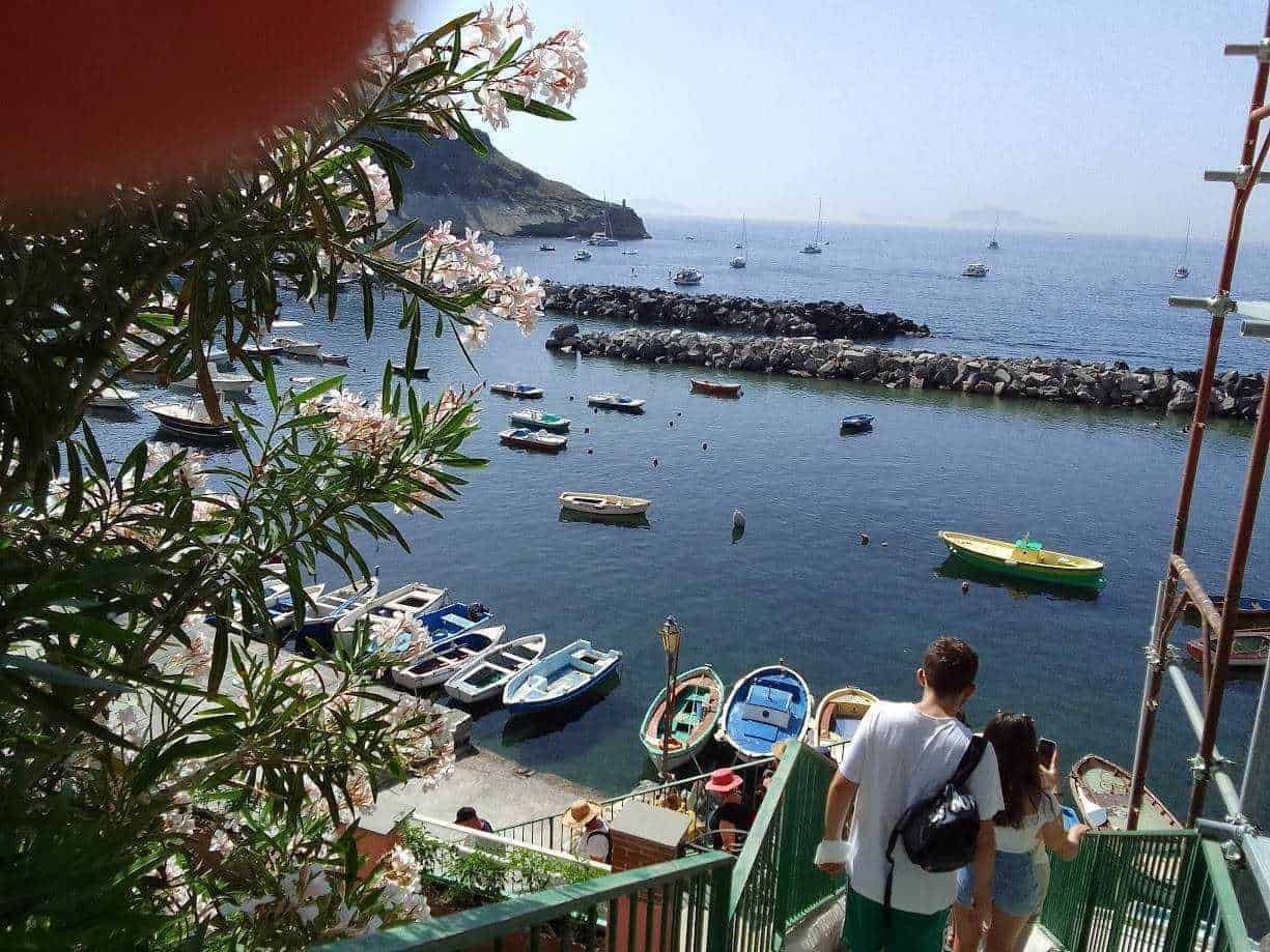 Barche nel porto di una località balneare con scogli, mare calmo e turistici che camminano lungo le scale. Primaverile e turistica, con fiori e scenari marini suggestivi.
