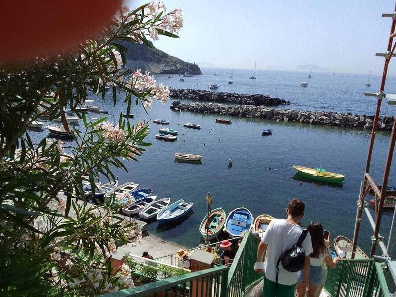 Procida: isola dei pescatori e cultura 12 Barche nel porto di una località balneare con scogli, mare calmo e turistici che camminano lungo le scale. Primaverile e turistica, con fiori e scenari marini suggestivi.