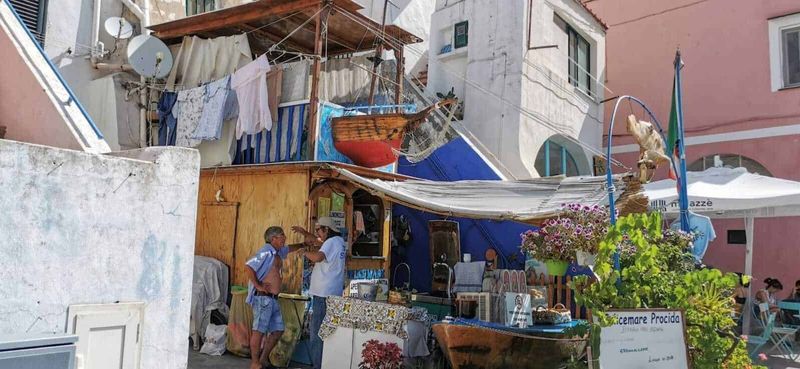 Procida: isola dei pescatori e cultura 13 Vicolo colorato con bancarella di street food e persone che conversano, case pittoresche con facciate in tonalità pastello, panni stesi, e decorazioni floreali in un quartiere caratteristico di pescatori.