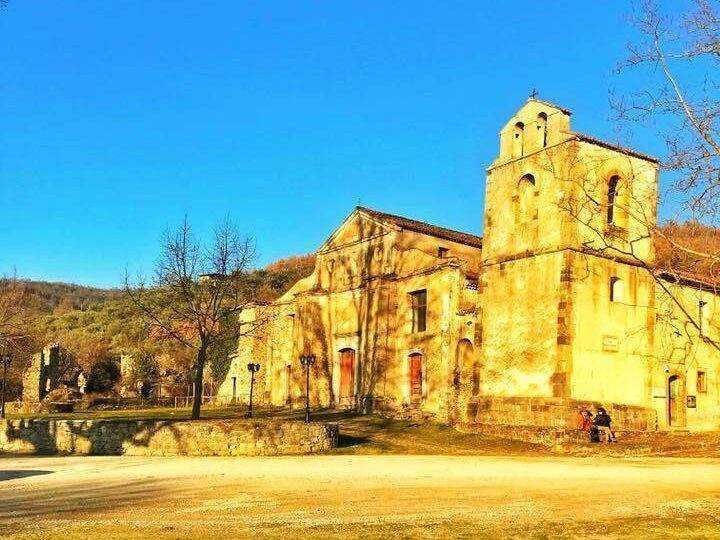 Vecchia chiesa in pietra con campanile in stile storico, situata in un paesaggio collinare con alberi e vigneti autunnali, ambientazione suggestiva e ricca di fascino culturale.