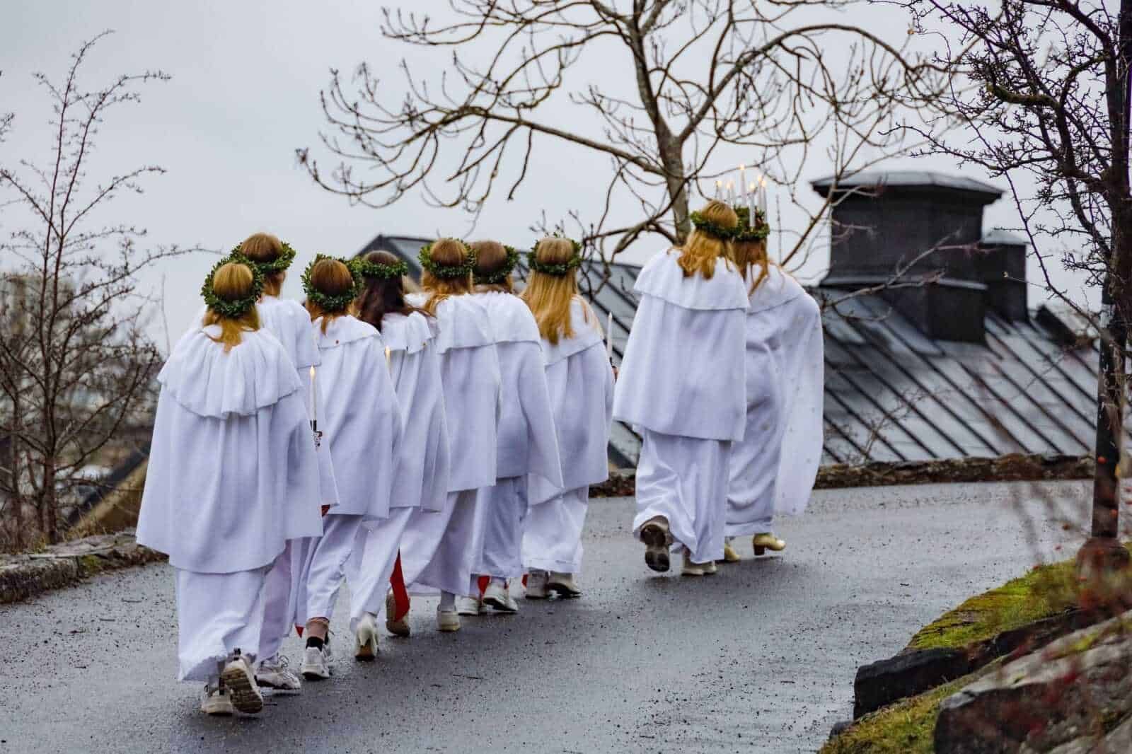 Preziosi angeli vestiti di bianco con corone di foglie sul capo, camminano su un sentiero di città con cielo nuvoloso. È una rappresentazione di un rito religioso tradizionale natalizio.