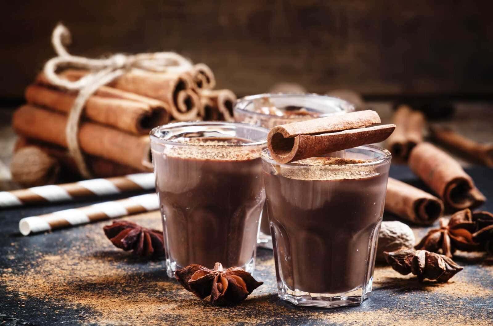 Deliziose cioccolate calde con cannella in bicchieri di vetro, accompagnate da bastoncini di cannella e spezie, perfette per le idee di ricette e dolci autunnali.