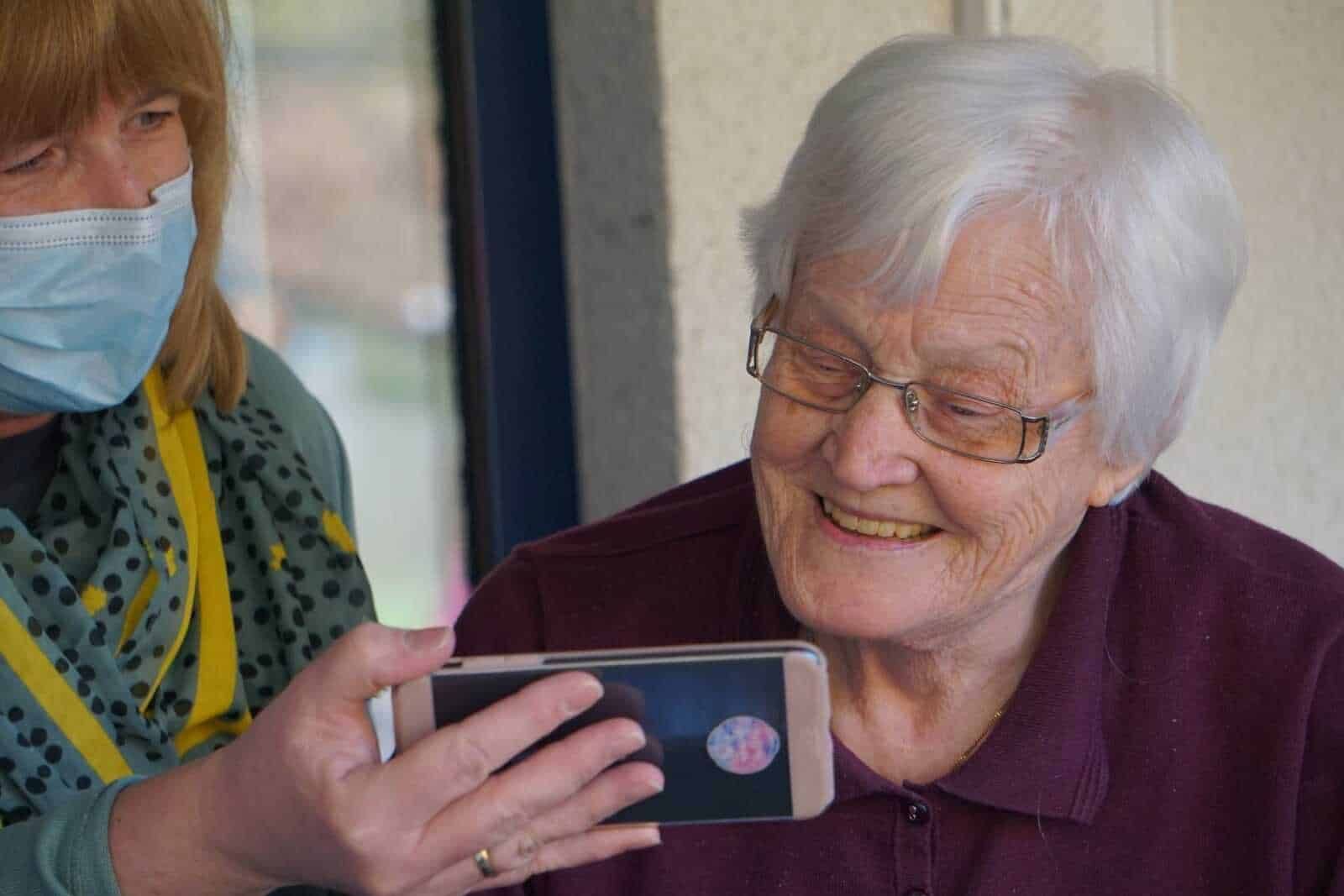 Vecchia donna sorridente che guarda il telefono di un'assistente con mascherina, in ambienti pubblici in Italia.