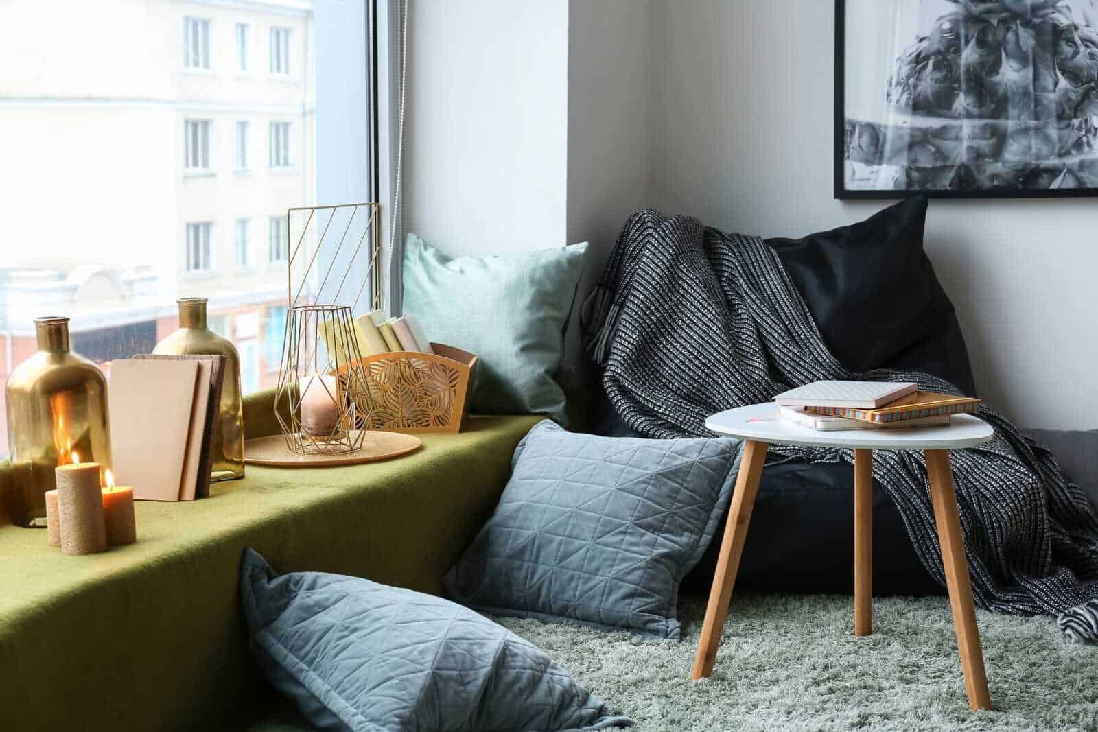 Soggiorno moderno con pouf grigio, candele accese, libri e decorazioni in stile minimalista vicino alla finestra luminosa e alle pareti chiare.