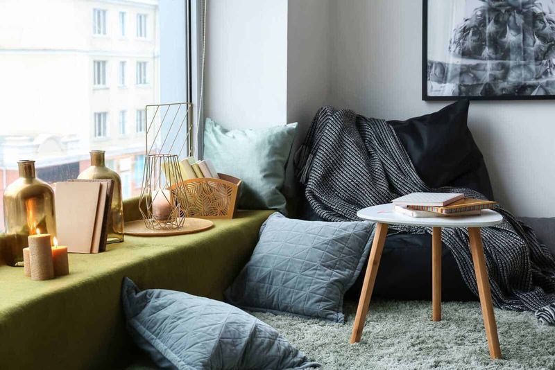 Arredare casa in stile hygge in 10 step 5 Soggiorno moderno con pouf grigio, candele accese, libri e decorazioni in stile minimalista vicino alla finestra luminosa e alle pareti chiare.