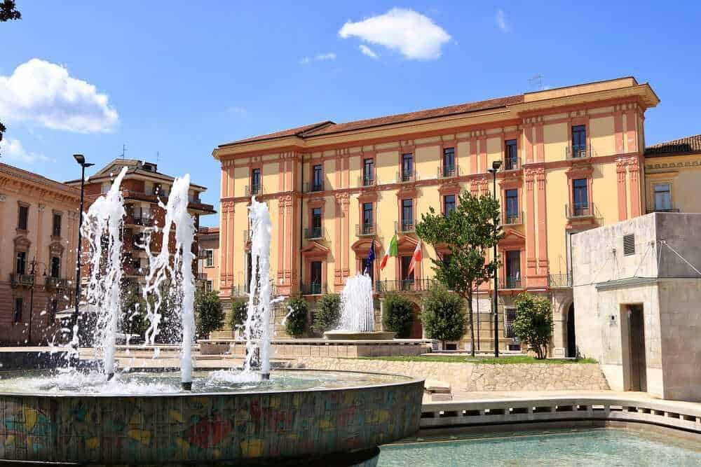 Faro di Fontana decorativa in una piazza urbana con edificio storico sullo sfondo, cielo blu e vegetazione, ambiente di città italiana, ideale per immagini di turismo e architettura.