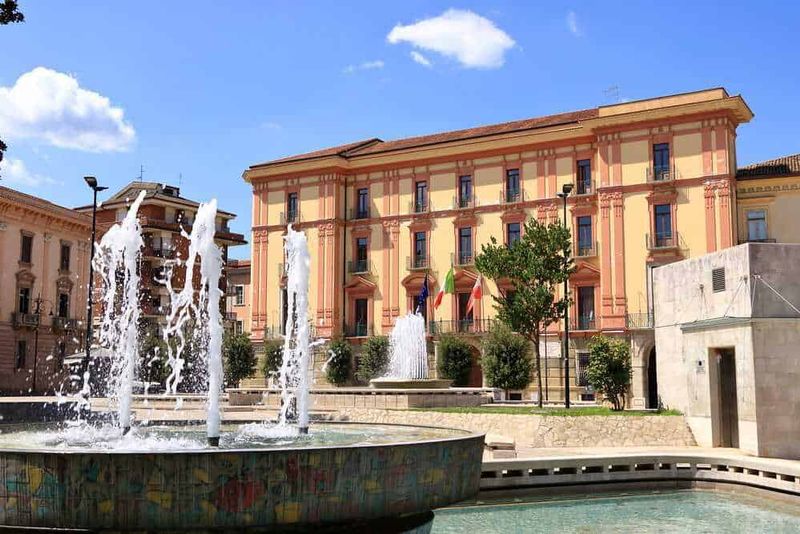 Luoghi da visitare ad Avellino e dintorni! 2 Faro di Fontana decorativa in una piazza urbana con edificio storico sullo sfondo, cielo blu e vegetazione, ambiente di città italiana, ideale per immagini di turismo e architettura.