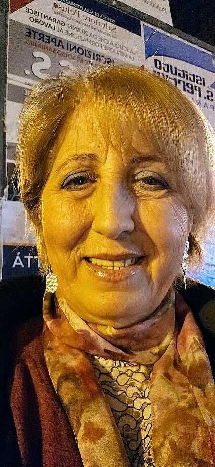 Una donna sorridente in posa davanti a uno sfondo di manifesti e pubblicità, indossa orecchini pendenti e una sciarpa colorata, perfetta per contenuti di blog su eventi, lifestyle e lifestyle femminile.