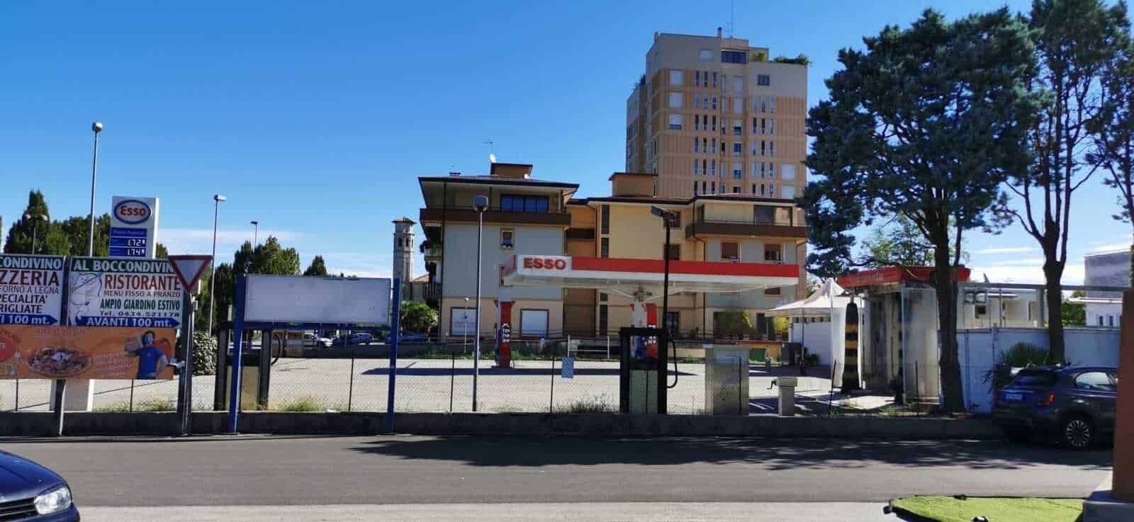 Esso stazione di servizio con edificio residenziale e spazi commerciali circostanti, nel contesto urbano, alto cielo azzurro, alberi e parcheggio adiacente.