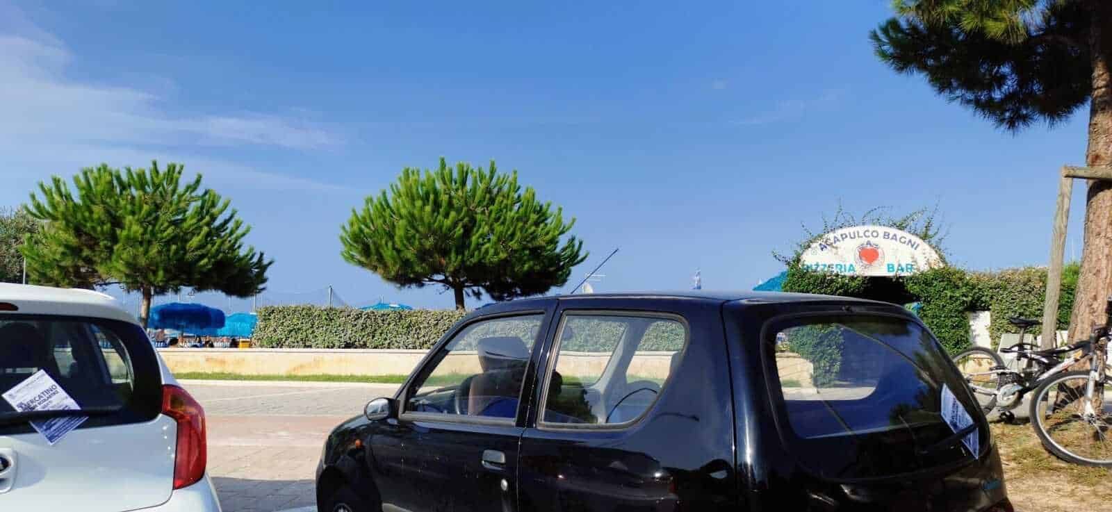 Auto d'epoca in un parcheggio vicino al mare con pini e bar in background, perfetto esempio di atmosfera rilassante in località balneare italiana.
