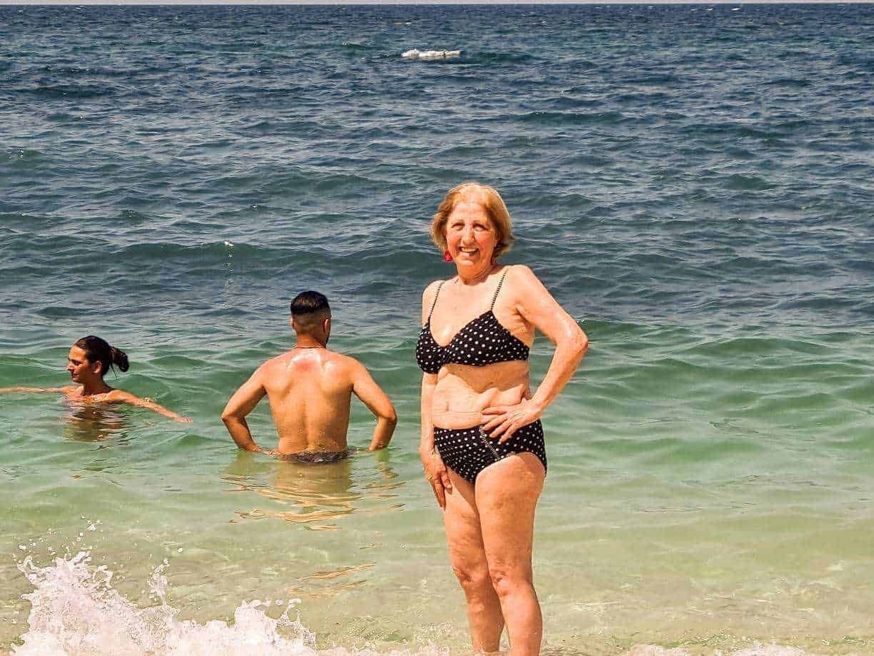 Vecchia donna in costume a pois sulla spiaggia del mare, sorridente e felice, tra turisti in acqua. Scene di relax e divertimento estivo sulla spiaggia mediterranea.