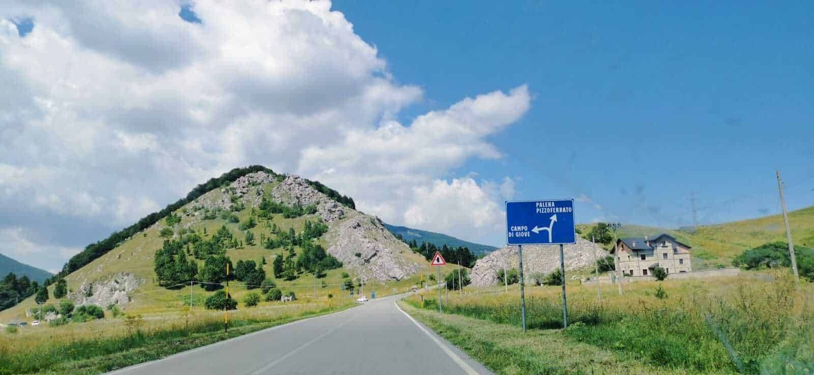 Montagna verde con strada di campagna e segnaletica stradale in Italia, paesaggio naturale e cielo azzurro.