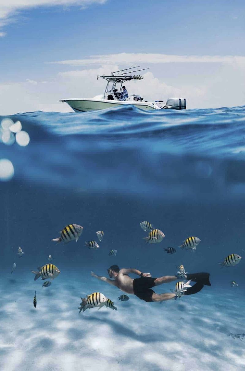 Avventure Subacquee: I Migliori Siti di Immersione 3 Remote boat on the sea with a diver swimming among colorful fish underwater, sunny sky, and ocean horizon, illustrating marine exploration, fishing trips, or water sports.