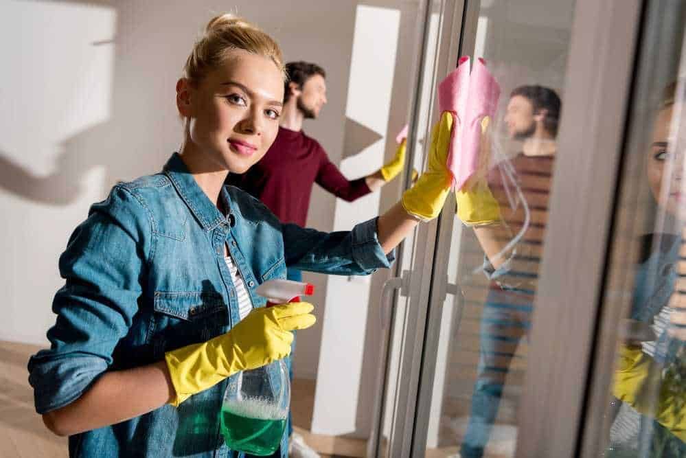Pulizie di vetri professionali con candeggina e panno rosa, donna sorridente con guanti gialli, ambienti domestici, servizi di pulizia approfonditi, cura della casa.