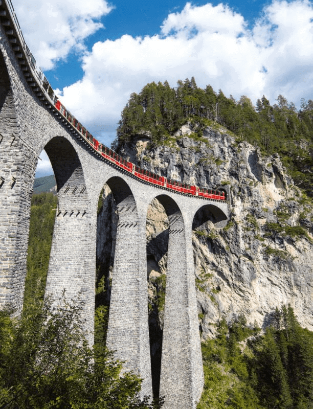 Ponti in pietra sopra una vallata montana con treno rosso in corsa, paesaggio naturale di alte montagne e cielo nuvoloso, esempio di ingegneria ferroviaria e scenari alpini.