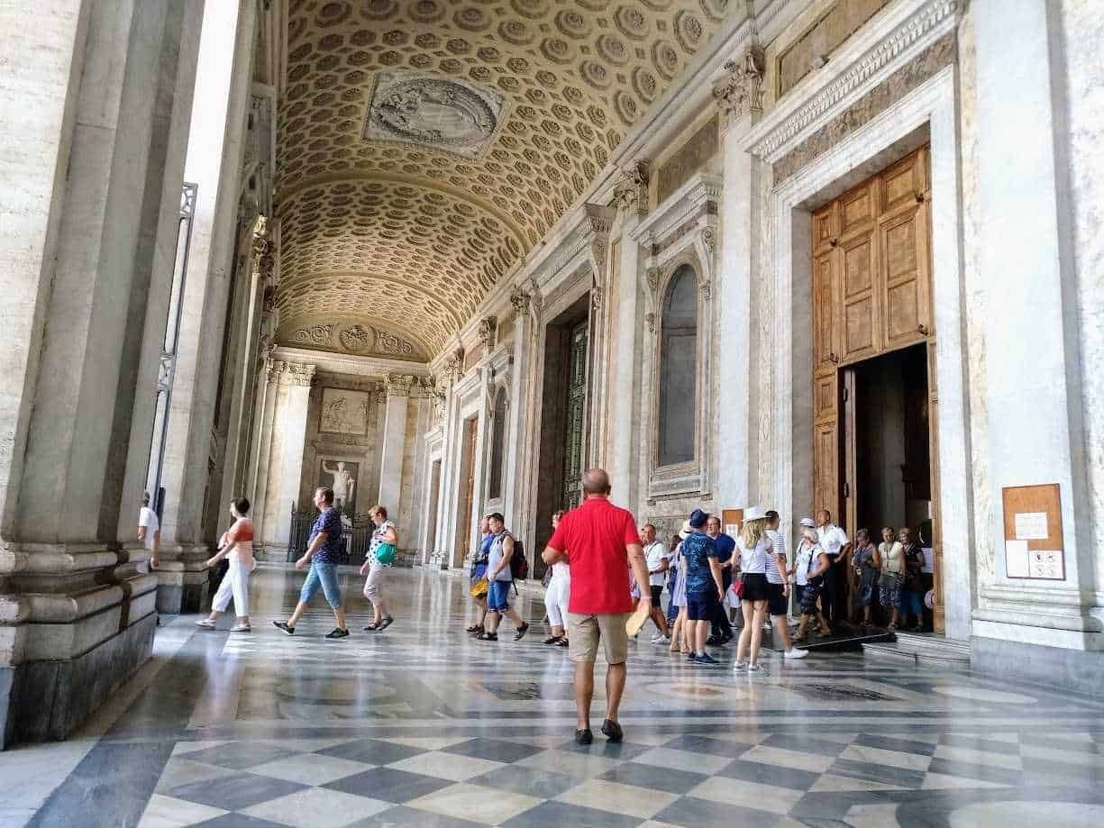 Immagine di un grande salone storico con soffitto decorato e pareti in marmo, visitato da un gruppo di turisti che osservano le opere d’arte e l’architettura elegante, perfetta per appunti sul blog di viaggi e cultura.