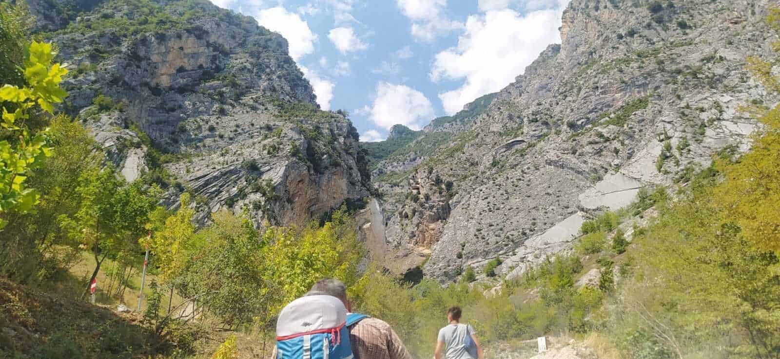 Escursionisti in montagna con zaini che attraversano una valle alpina circondata da scoscese pareti rocciose e vegetazione verde, in un paesaggio naturale e suggestivo.