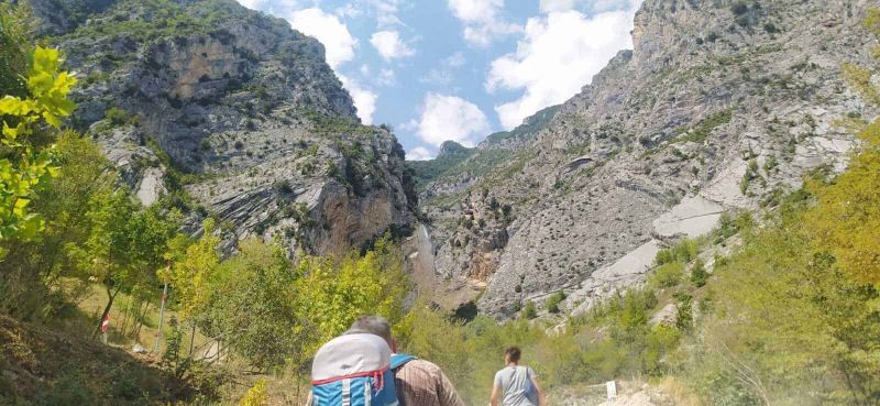 Esplorando il Fascino del Paesaggio di Montagna 5 Escursionisti in montagna con zaini che attraversano una valle alpina circondata da scoscese pareti rocciose e vegetazione verde, in un paesaggio naturale e suggestivo.