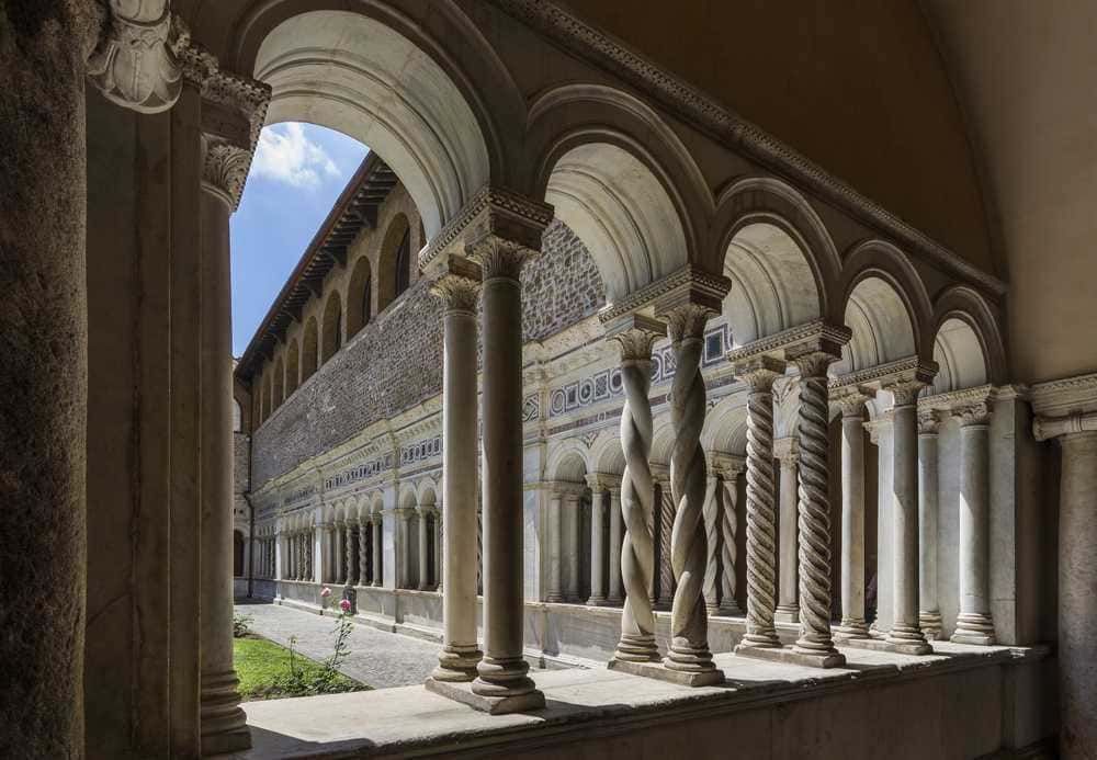 Antichi archi e colonne in marmo in un chiostro storico, esempio di architettura medievale con dettagli decorativi e stile rinascimentale. Spazio tranquillo e suggestivo con cielo azzurro visibile.