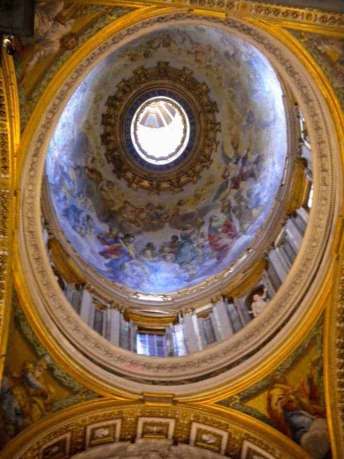 Opulenta cupola decorata con affreschi religiosi e dettagli dorati, illuminata dalla luce naturale che penetra attraverso un oculo centrale, creando un'atmosfera di grandiosità e spiritualità.
