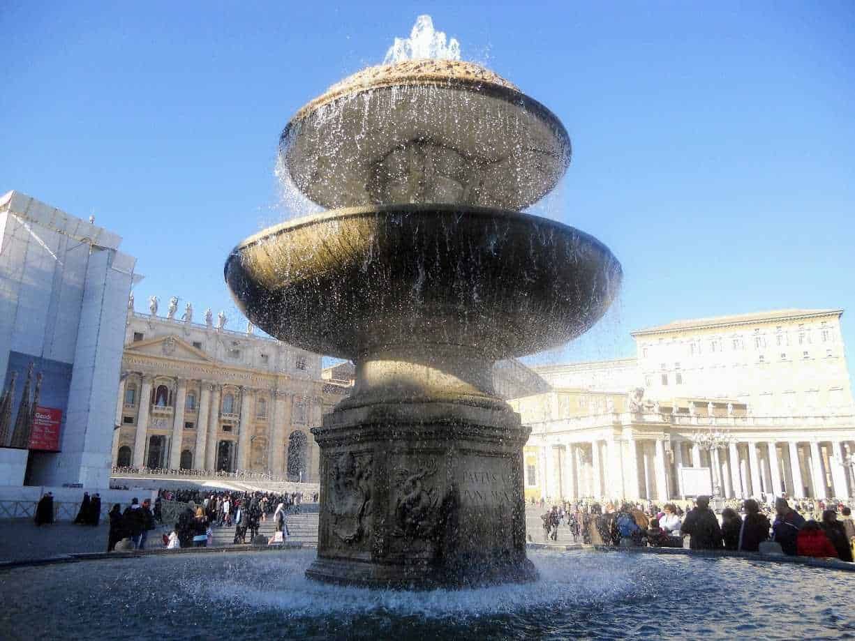 Fontana storica al centro di Piazza San Pietro, con architettura barocca e fontana in pietra, sotto un cielo blu, affollata di visitatori in un giorno di sole.