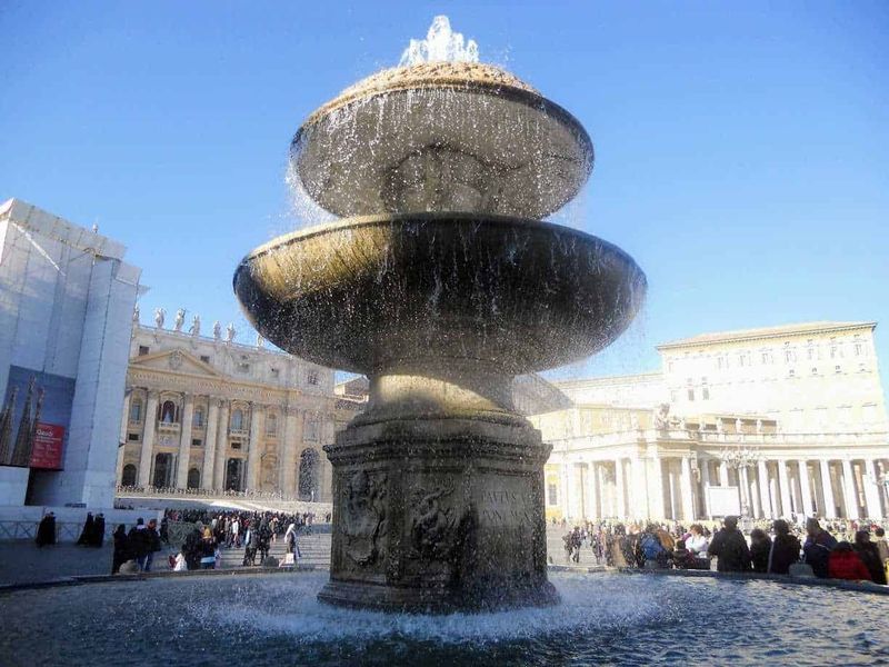 7 Cose da vedere in Piazza San Pietro gratis 11 Fontana storica al centro di Piazza San Pietro, con architettura barocca e fontana in pietra, sotto un cielo blu, affollata di visitatori in un giorno di sole.