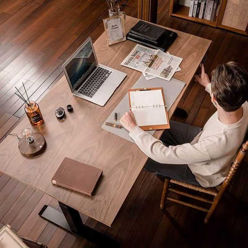 Laptop open con schermo acceso, quaderno aperto, penna, giornale e libri sulla scrivania in un ufficio casalingo.