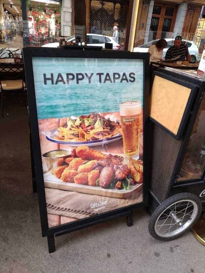 Le tapas: un'autentica esperienza culinaria made in Spain 3 Bacheca pubblicitaria con immagine di tapas e birra, promozione di Happy Tapas in un ristorante con clienti seduti al tavolo.