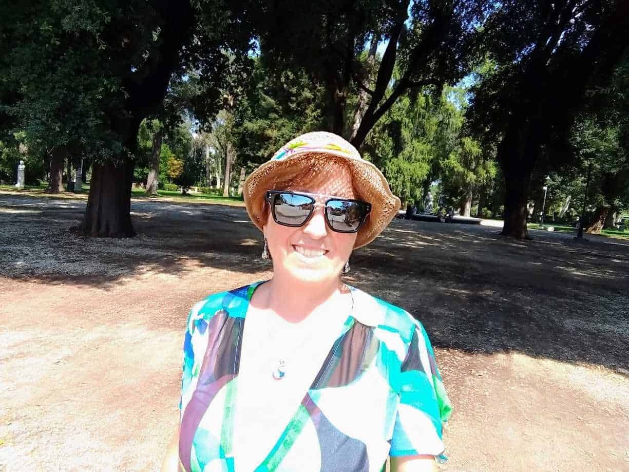 Fotografia di una donna sorridente con cappello di paglia e occhiali da sole, passeggiata in un parco con alberi verdi e ombreggiati, atmosfera estiva e rilassante.