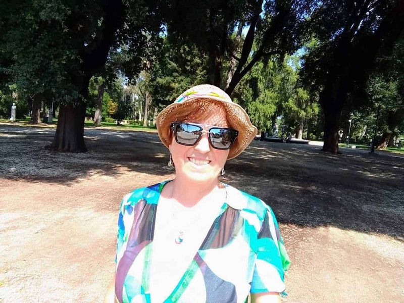 Scopri l’unico orologio idraulico di Roma 6 Fotografia di una donna sorridente con cappello di paglia e occhiali da sole, passeggiata in un parco con alberi verdi e ombreggiati, atmosfera estiva e rilassante.