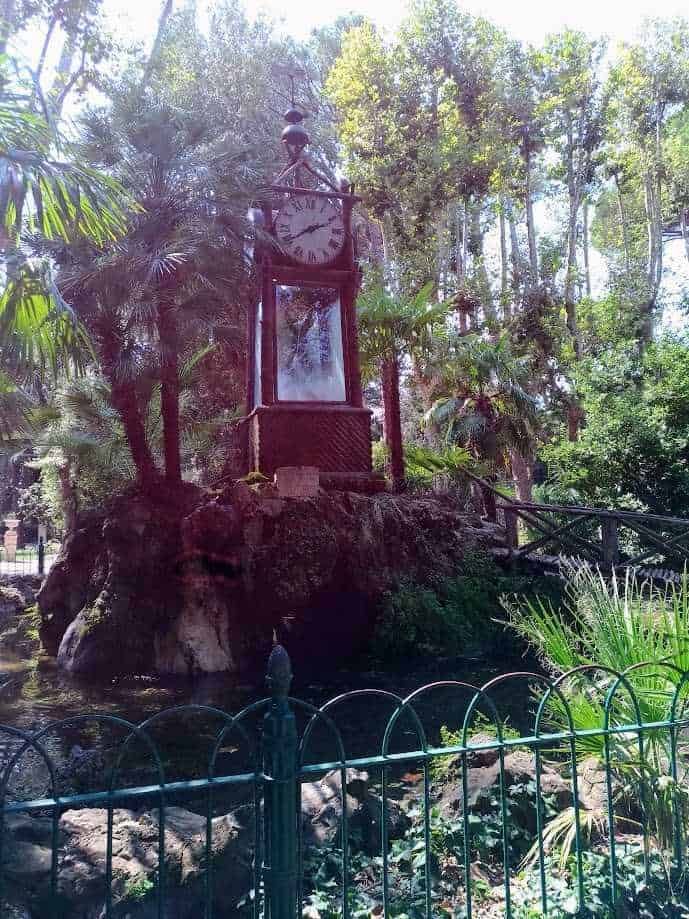 Vecchio orologio da torre in stile vintage situato in un parco verde con alberi e piante tropicali, circondato da una recinzione di ferro, in un ambiente naturale e rigoglioso.