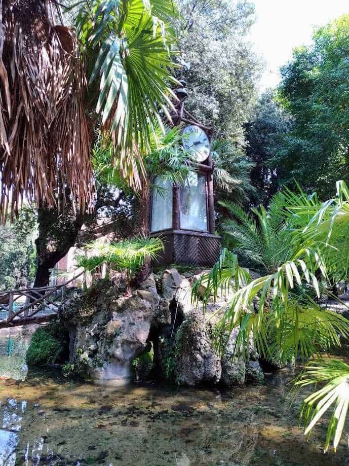Clock da giardino in stile vintage immerso in una rigogliosa vegetazione tropicale, circondato da piante verdi e rocce, creando un'atmosfera rilassante e naturale in un giardino esotico.