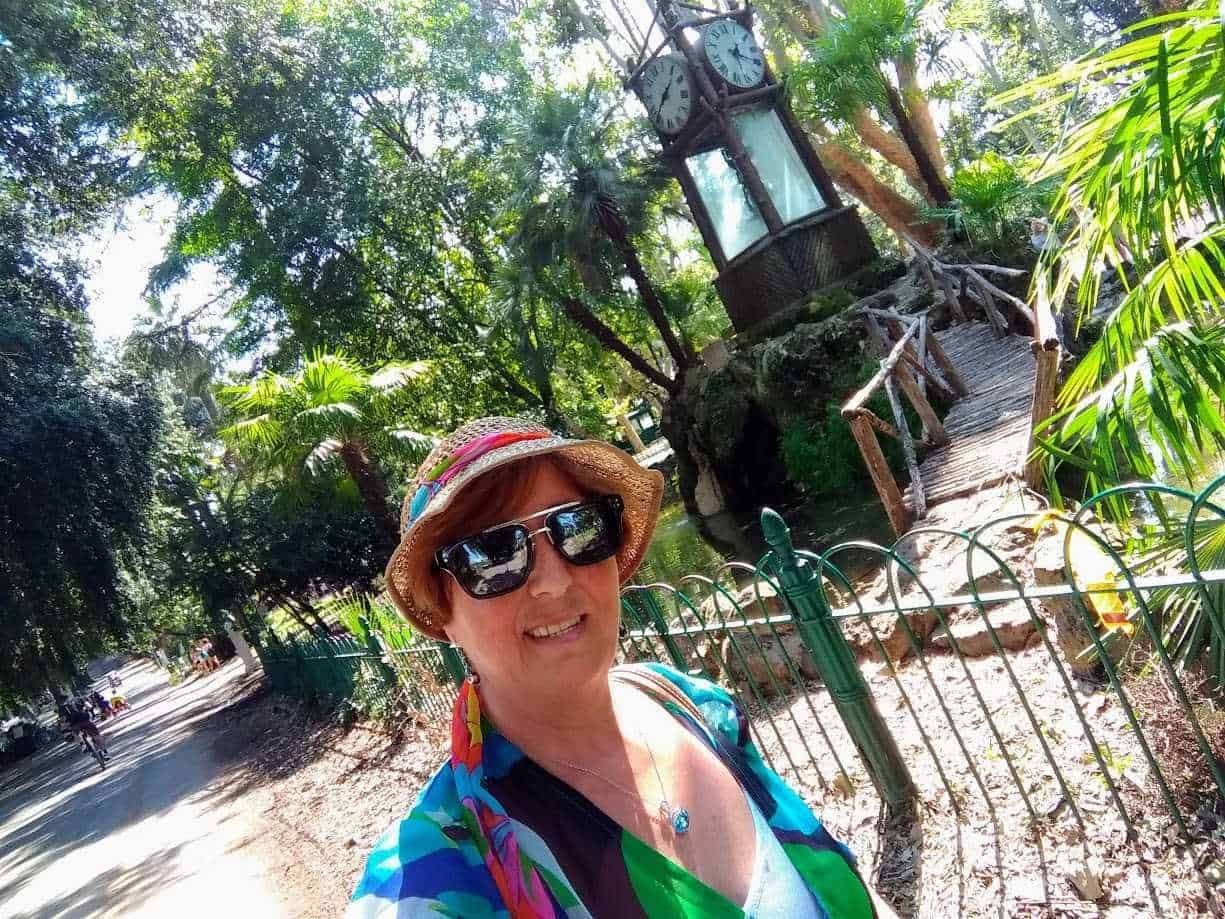 Vecchietto in un parco con orologio di vetro e tetto di paglia, sorridente con cappello e occhiali da sole alla moda, in un ambiente verde lussureggiante, perfetto per esplorare la natura e scattare foto ricordo.