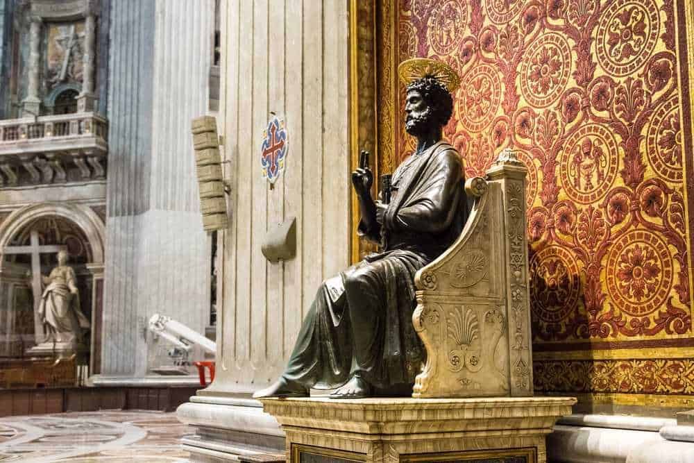 Statuetta di San Paolo in bronzo all'interno di una chiesa storica con dettagli decorativi dorati e pareti con tessitura elaborata, simbolo di fede e arte sacra.