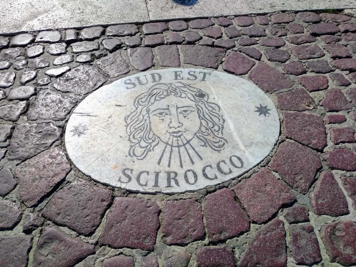 Sud Est Scirocco manhole cover su strada cittadina, elemento di design urbano con decorazioni artistiche e testi.
