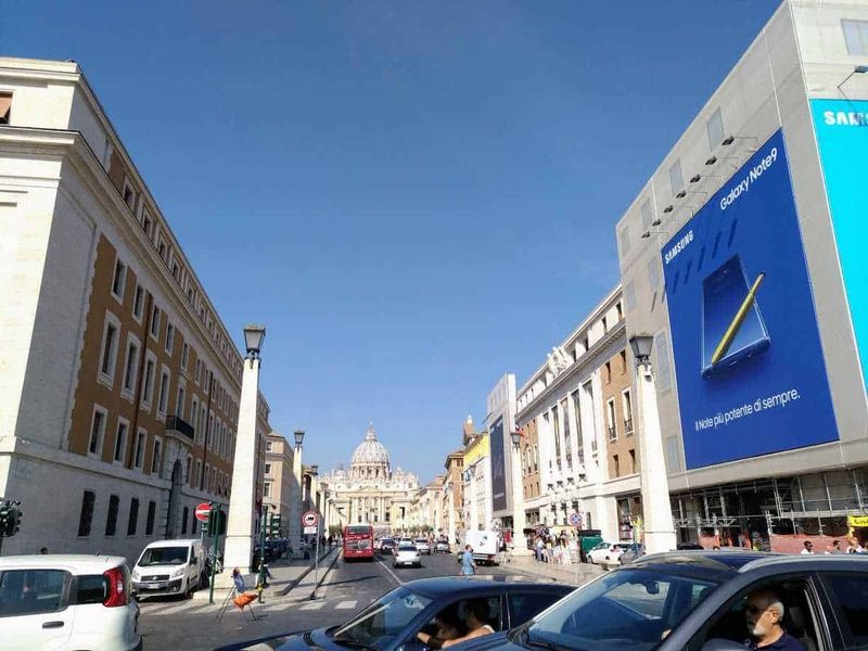 7 Cose da vedere in Piazza San Pietro gratis 13 Samsung Galaxy Note9 pubblicità gigante su edificio storico a Roma, vista strada trafficata con cittadini e auto, alla scoperta di luoghi iconici della città eterna.