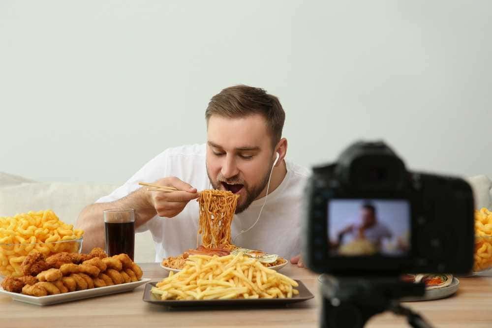 Sorrento di spaghetti su un piatto, uomo che mangia con forchetta durante la registrazione di un video food blog, con vari piatti di cibo e una videocamera davanti, ambientazione casalinga.