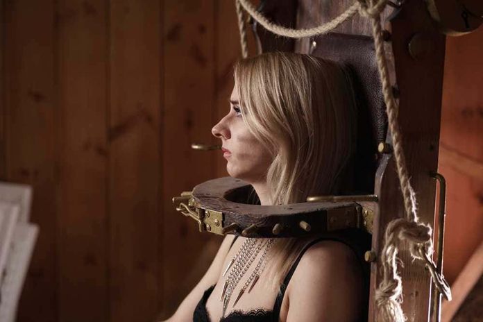 15 cacciatori di streghe più famosi nella storia 5 Collare di tortura medievale con donna seduta, immagine storica, ambiente in legno, versione moderna e originale, tema di torture e storia medievale, rappresentazione di tortura e sofferenza, contenuti storici e di interesse culturale, immagine di prova di resistenza e tormento.