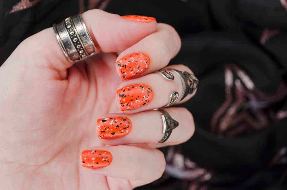 Unghie corte autunno 2024: Effetto manicure con smalto arancione con spruzzi neri e argento, mani curate con anelli alla moda, focus su unghie e stile glamour.