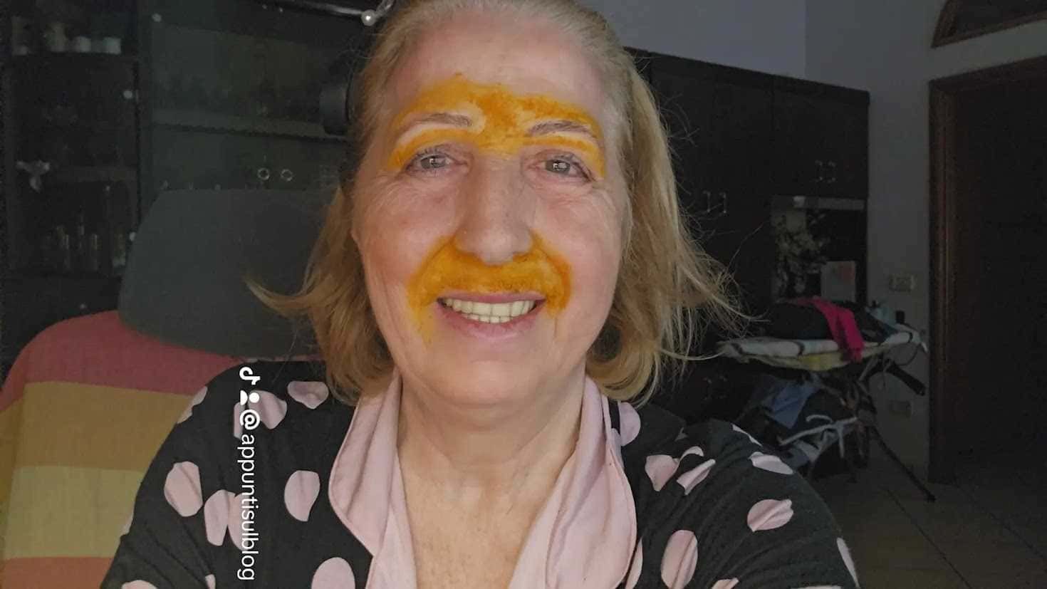 Ragazza sorridente con trucco originale di colore arancione sulla fronte e sul naso, in un ambiente domestico, ritratta mentre fa un selfie in casa.