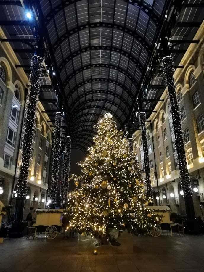 Albero di Natale decorato con luci e ornamenti dorati sotto una volta in vetro laccato, con colonne illuminate, atmosfera natalizia elegante in un atrio storico.